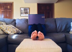 Person on a sofa with face hidden behind a laptop; only their feet are visible. Working at home.