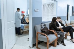 physician and patient in consulting room with patients waiting outside on hospital chairs